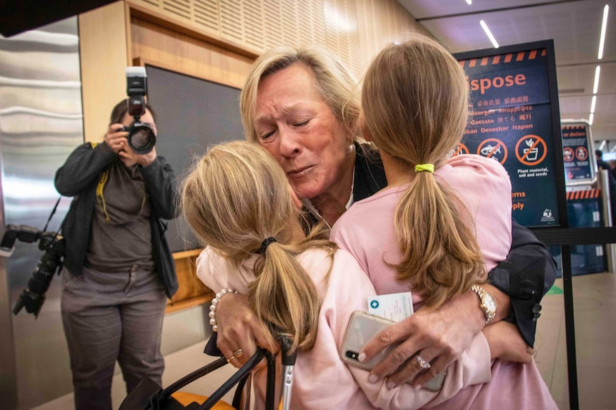 A woman hugs two young girls at an airport