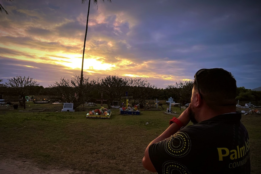 A man looks out towards graves with palm trees and a cloudy sunset in background. 