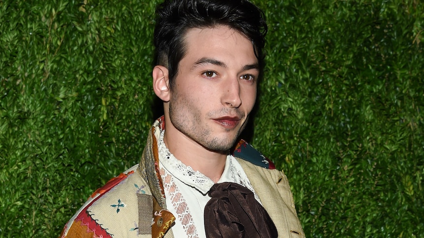 A person with short black hair in front of brown backdrop, wearing dark lipstick and wearing jacket