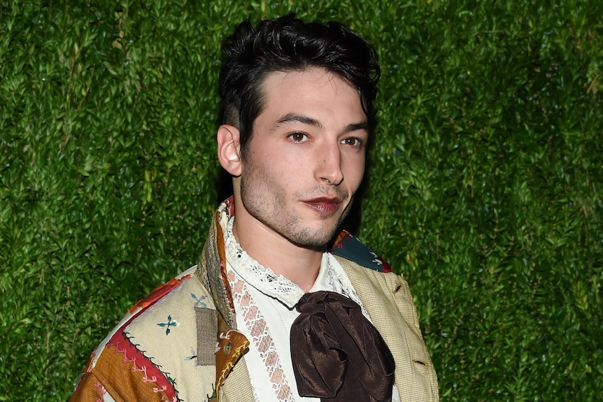 A person with short black hair in front of brown backdrop, wearing dark lipstick and wearing jacket