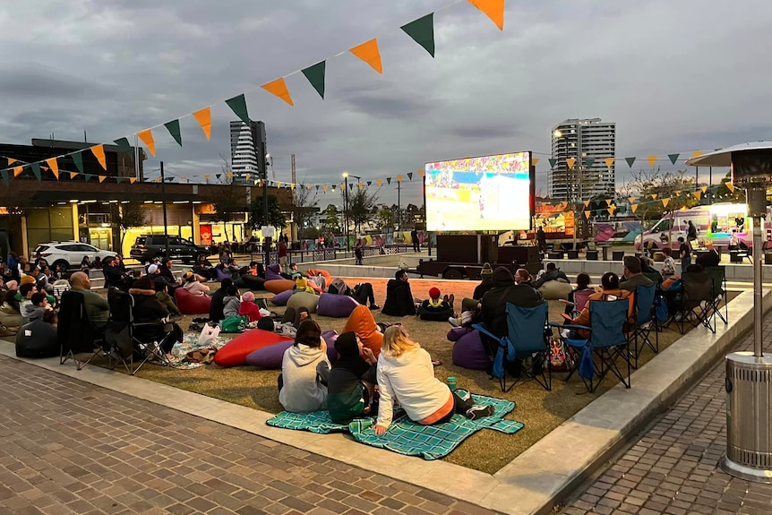 Crowds of football fans watch a large screen outside displaying a football match.