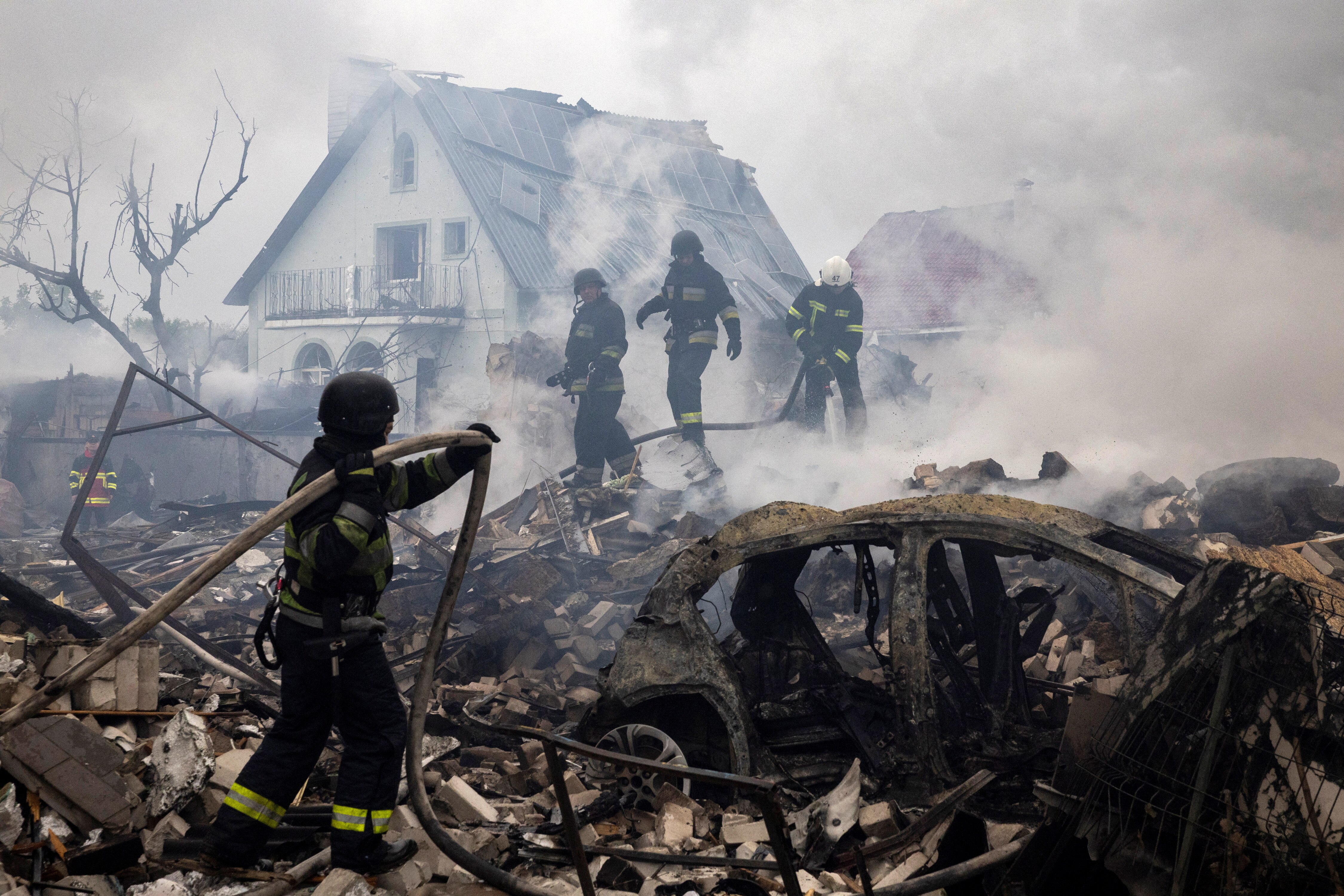 Firefighters work amid smoke, rubble, and a destroyed car, with a damaged house visible in the background.