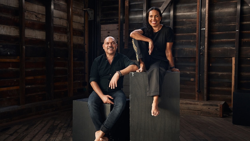 An Indigenous man in his mid 50s and an Indigenous woman in her late 40s sitting on black boxes in a wooden room