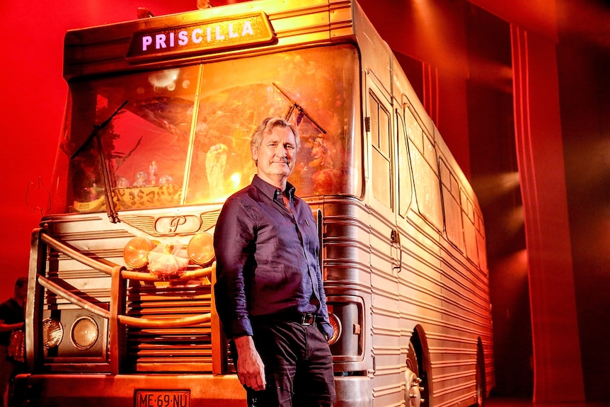 An older white man posing in front of a big gold bus called 'Prisciila'