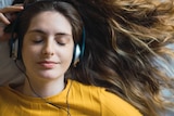 A portrait of a smiling young woman lying on a bed listening to something on her headphones.