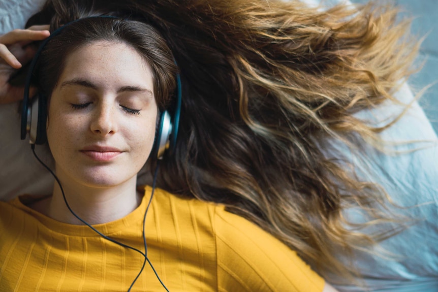 A portrait of a smiling young woman lying on a bed listening to something on her headphones.