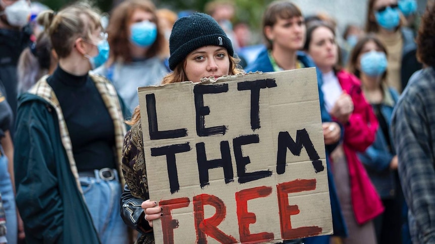 A young woman wearing a beanie holds a sign 'Let them free' at a protest at Kangaroo Point.