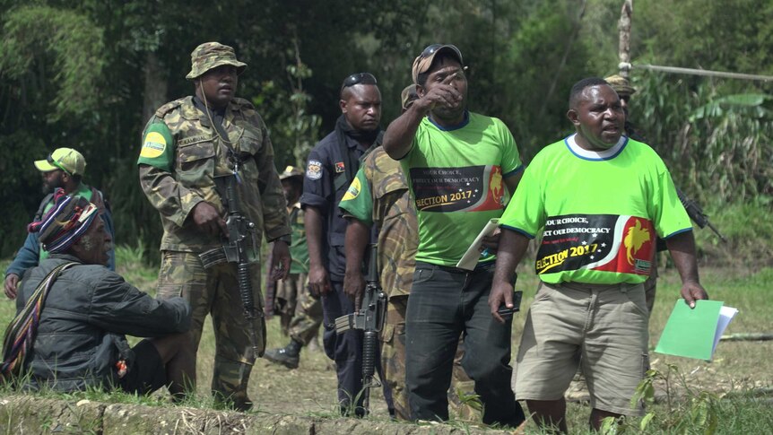 Two polling officials point to something out of shot as armed soldiers and police officers look on.