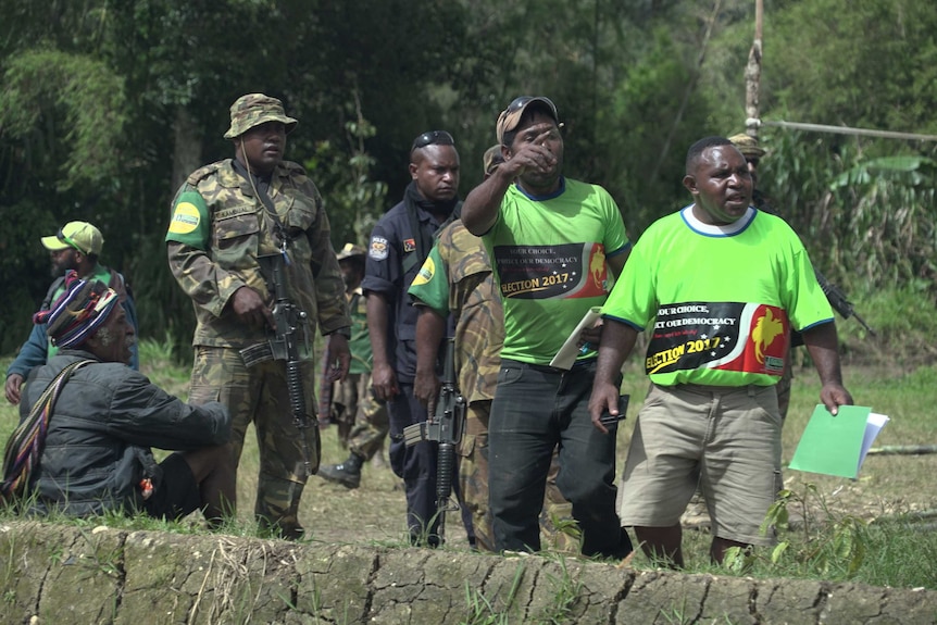 Two polling officials point to something out of shot as armed soldiers and police officers look on.