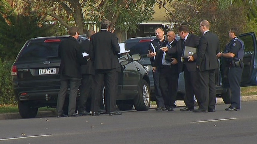 Police and people in suits stand and talk near a car.