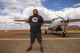 A young Aboriginal man standing on an outback runway in front of a small aircraft and looking serious.