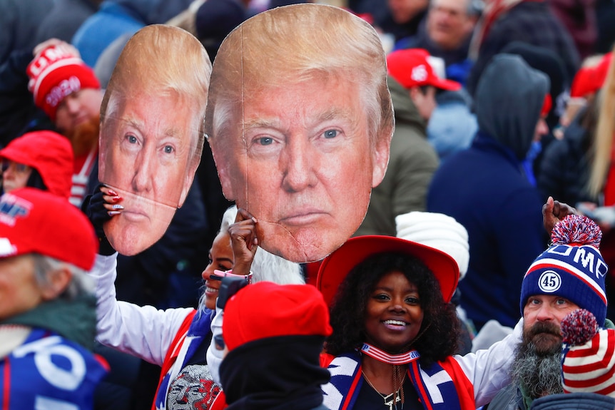 Two women hold up large cardboard cutouts of Donald Trum's face at a rally for supporters.