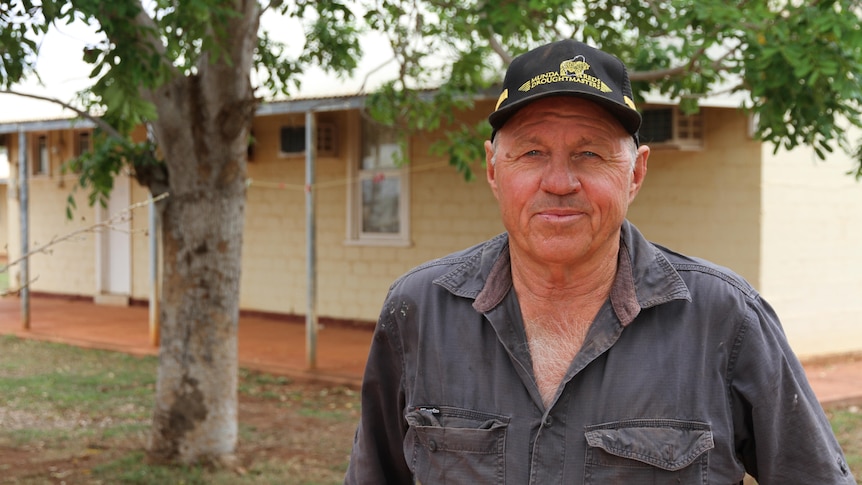 A man wearing a slate grey shirt and black cap with yellow writing, standing in front of a tree, looks at the camera