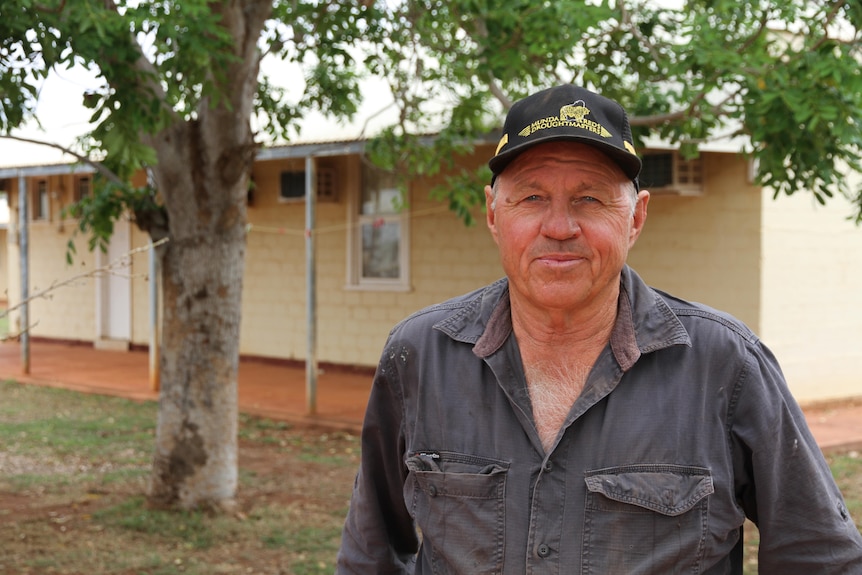 A man wearing a slate grey shirt and black cap with yellow writing, standing in front of a tree, looks at the camera