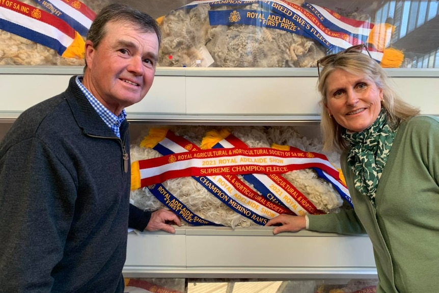 A man wearing a dark blue jumper and a lady in a green jumper stand in front of wool that has been shorn from a sheep. 