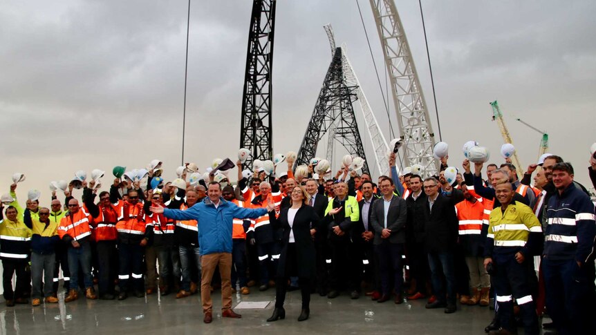 Workers in high-vis holding their hard hats aloft on a bridge with the WA Premier in the foreground.