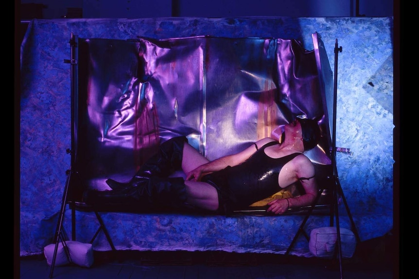 A man wearing black latex lies in a foil "trough", illuminated by blue light.