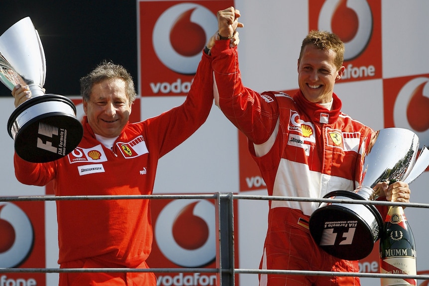 Two men in red racing suits, both holding winners trophies, raising their arms in victory.