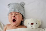 A small baby yawns while tucked into bed with a teddy bear.