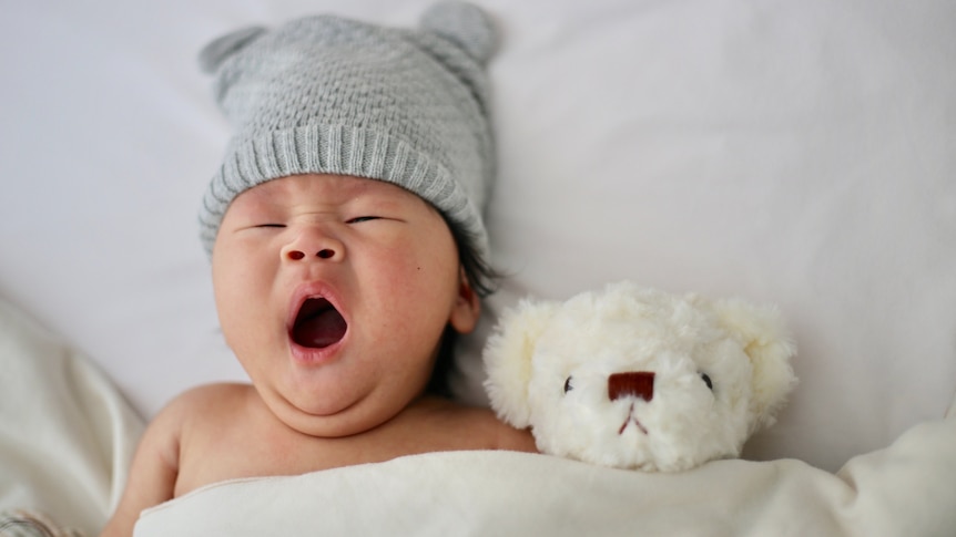 A small baby yawns while tucked into bed with a teddy bear.