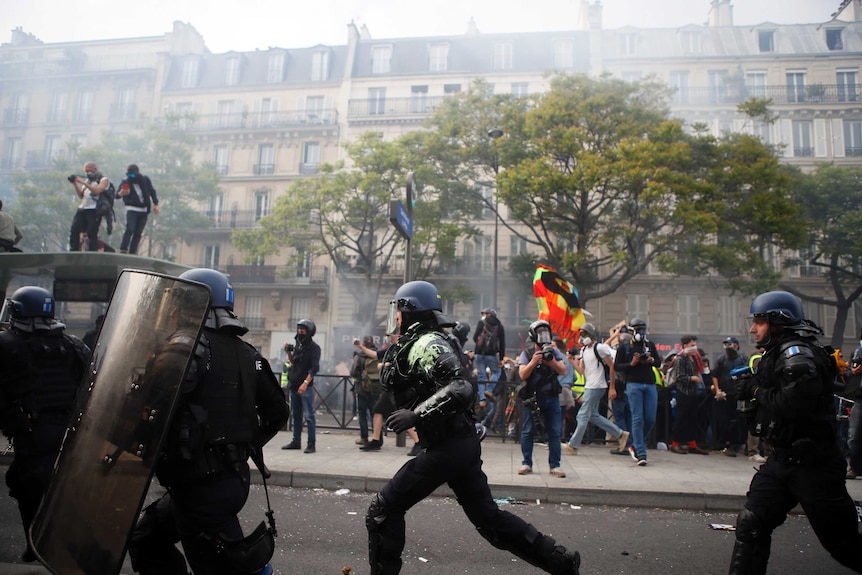 Police run in the street in riot gear and helmets with smoke from tear gas and protestors standing in the background.