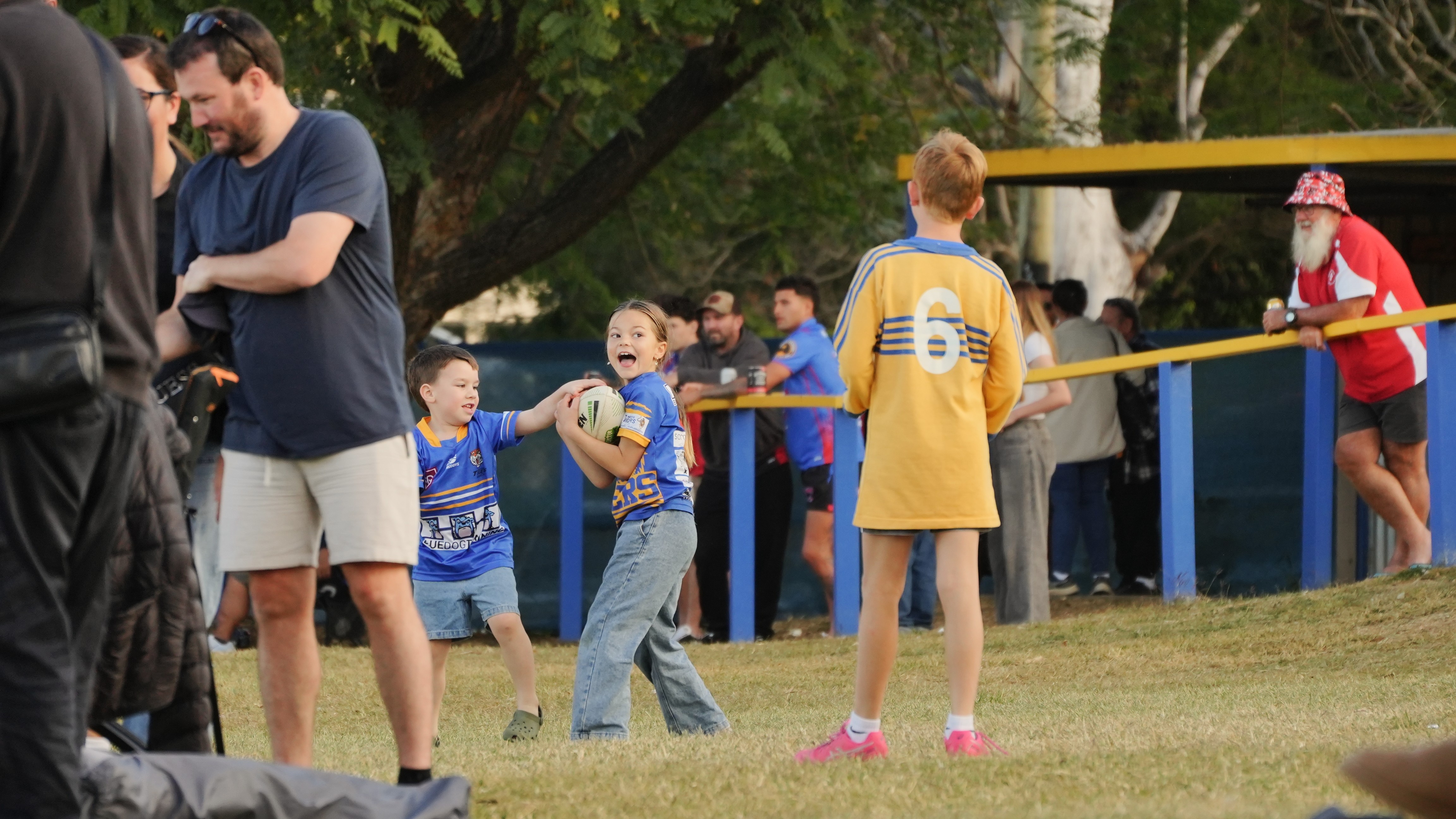 A young girl in a blue rugby jersey and jeans holding on to a ball with an excited look on her face.