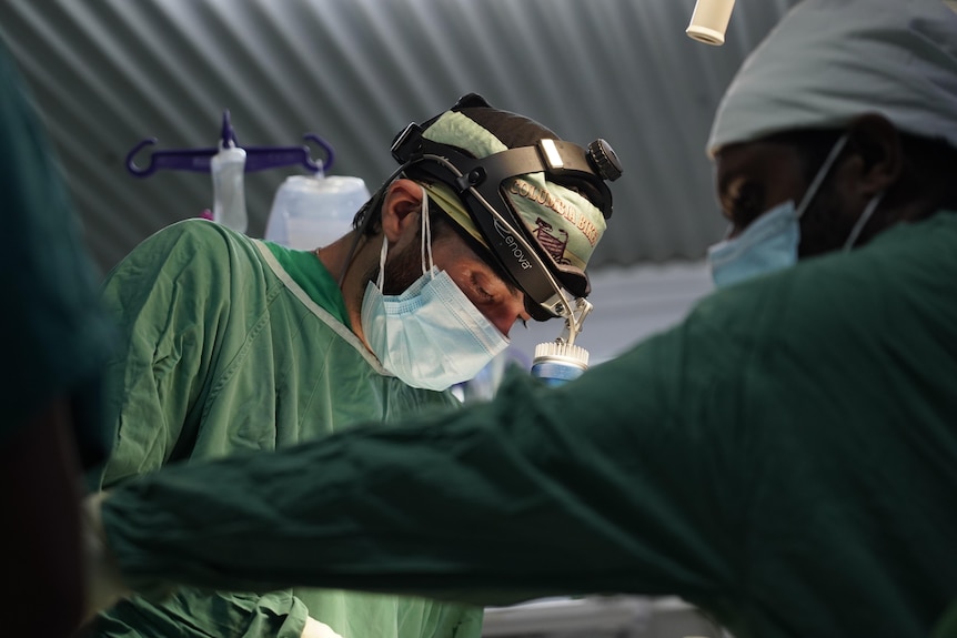 Two men wearing surgical attire, one with a head torch on top of his head, look down at a patient