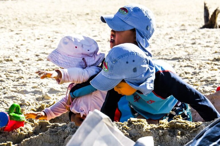 Three young kids on a beach wearing hats and pushing and pulling each other a toy