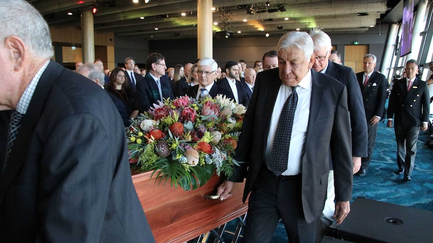 Suited men carry a coffin topped with native flowers in a crowded room.