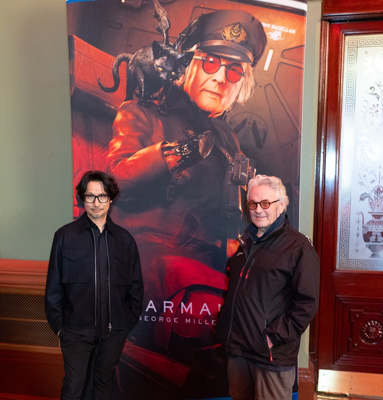 Hideo Kojima and George Miller in front of poster for Death Stranding with a character based on Miller