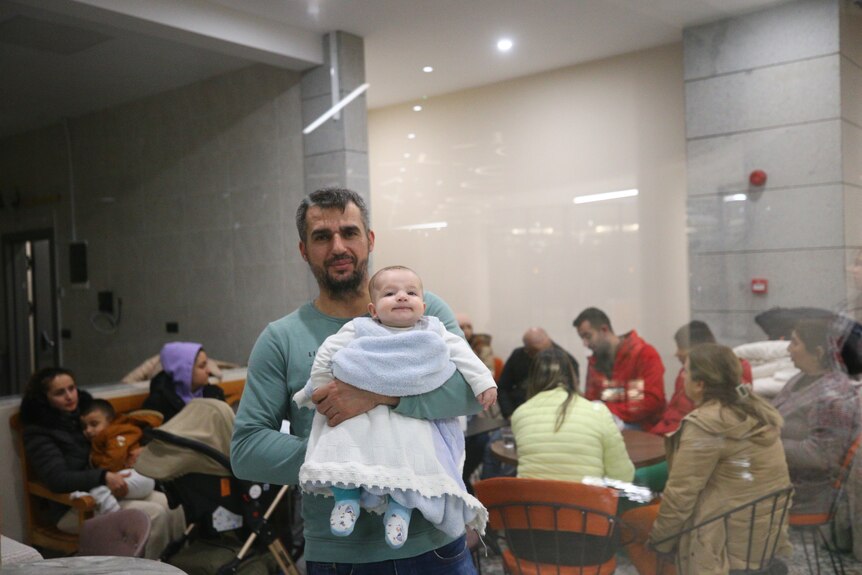 A man holds up a baby while people sit behind them. 