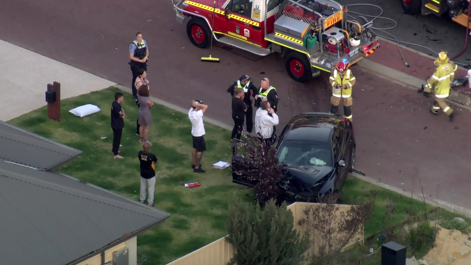 People stand on the front lawn of a house near a crashed black car, with police officers and firefighters nearby.