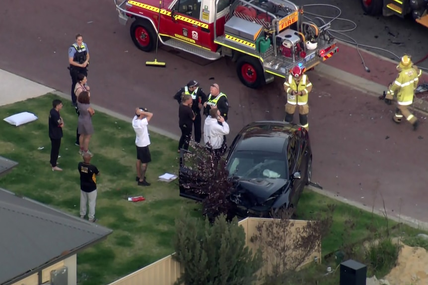 People stand on the front lawn of a house near a crashed black car, with police officers and firefighters nearby.