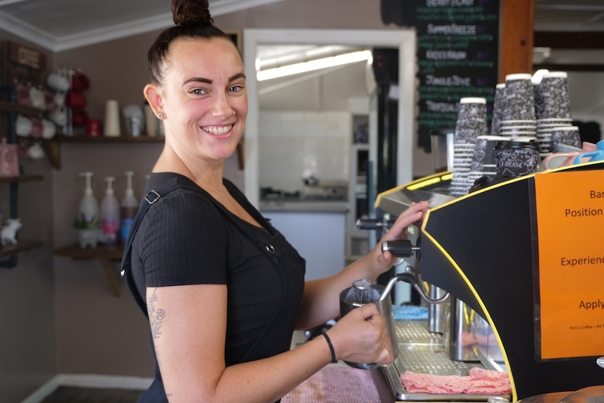 Sara Rowley, dark shirt, dark overalls, dark hair in a top knot, frothing milk in a jug at a coffee machine.