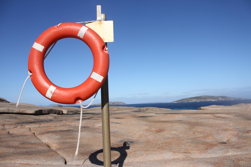 A round red life buoy is fixed to a pole on granite rocks by the ocean.