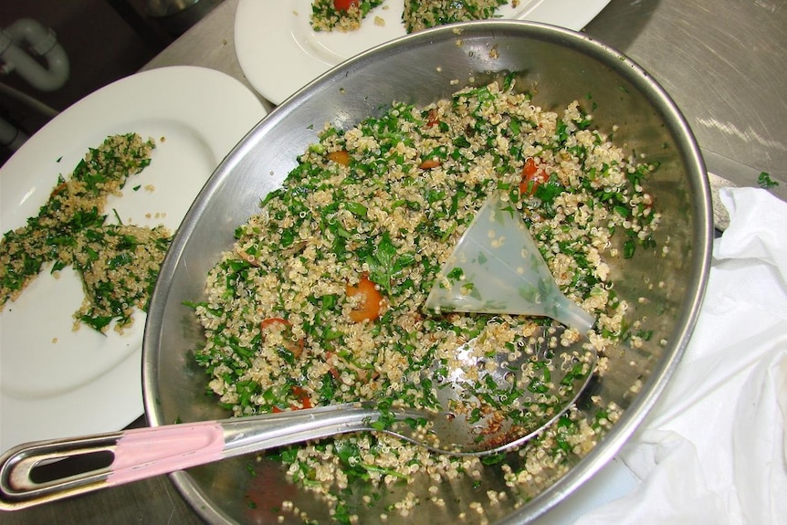 A large metal salad bowl and spoon with a salad made of pale grains, chopped tomato and parsley.