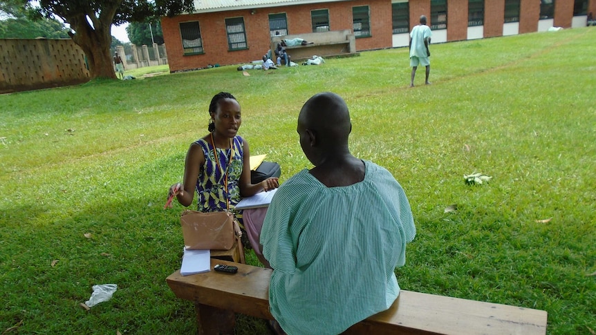African man with back to camera, in hospital smock, sitting with African woman, front to camera, taking notes