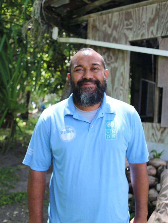 A smiling man wearing a pale blue polo shirt, bald, thick salt and pepper beard and moustache.