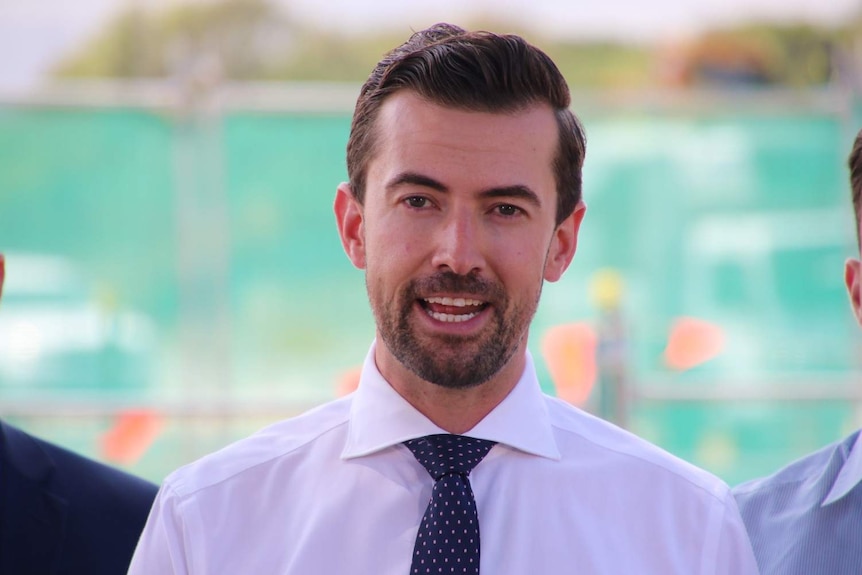 Zak Kirkup wearing a white shirt and blue spotted tie, speaking at a press conference.