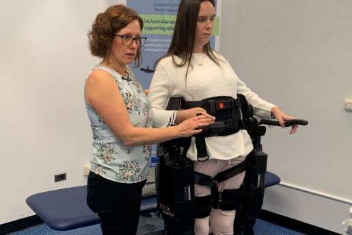 A young woman stands in a support device and another woman stands next to her supporting her.