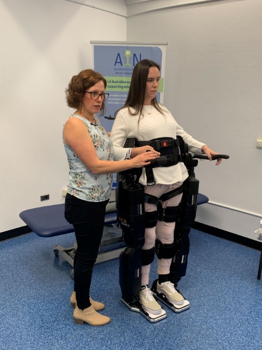 A young woman stands in a support device and another woman stands next to her supporting her.