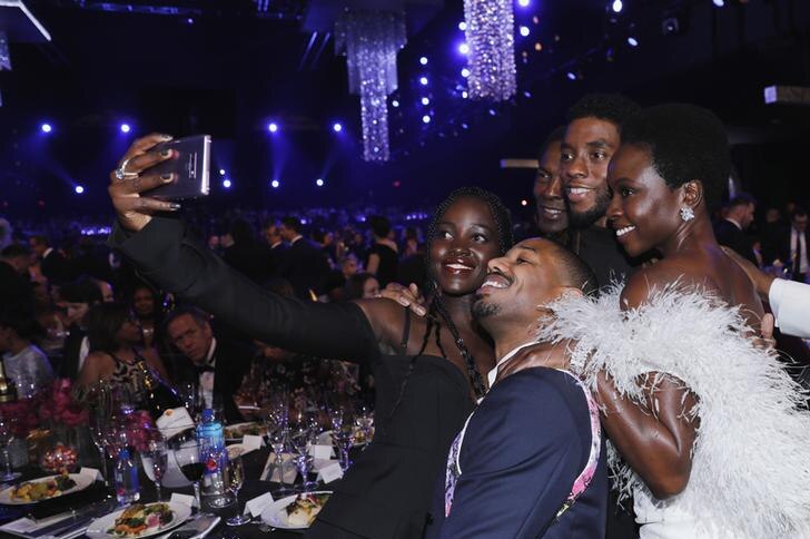 Danai Gurira grins at phone in white feather ballgown with hand on Michael B Jordan who squats near Lupita Nyong'o for selfie.