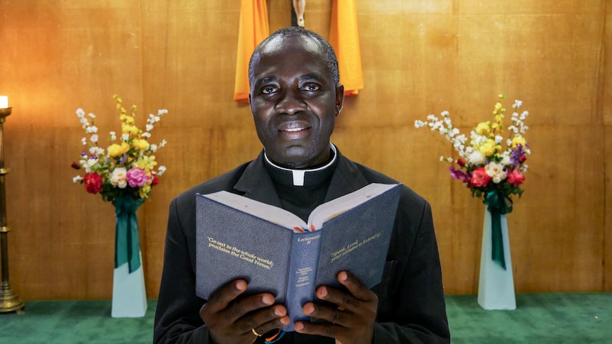 Father Emmanual Gyamfi stands in front of the cross with a bible.