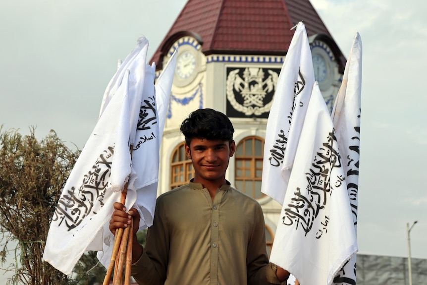 A teenaged boy stands in front of a mosque holding bunches of white and black Taliban flags