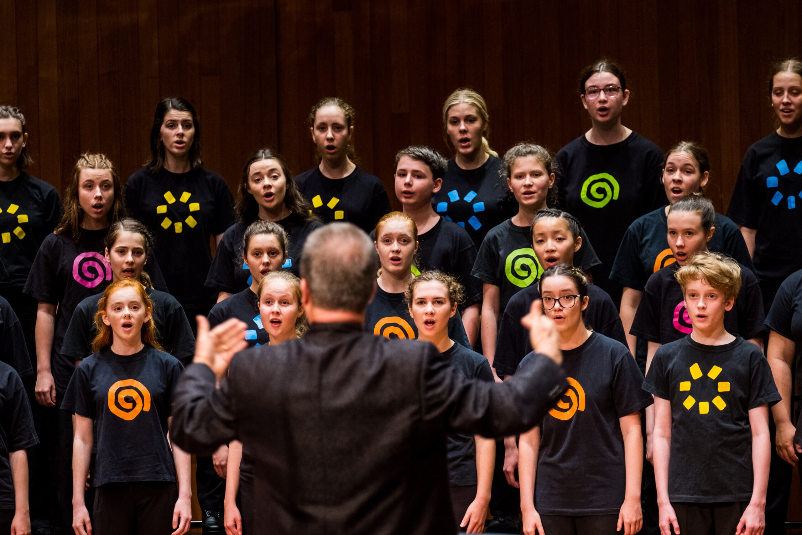 A man stands with his back to the camera conducting a children's choir. The choir wear black shirts with colourful designs.