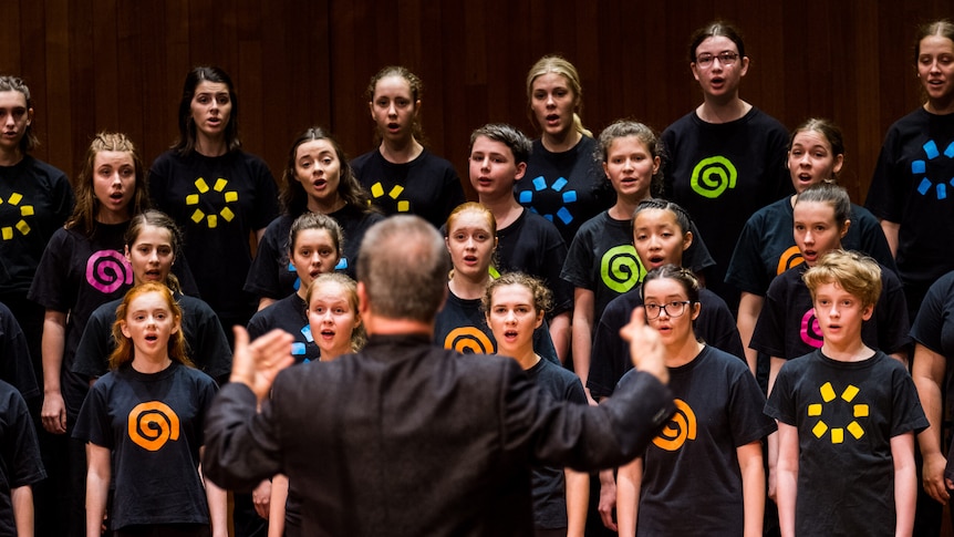 A man stands with his back to the camera conducting a children's choir. The choir wear black shirts with colourful designs.