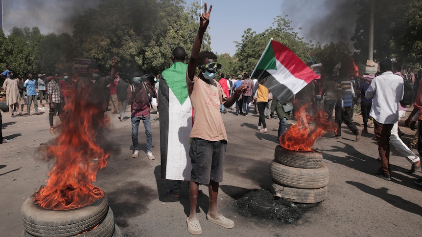 Man holds arm up with peace sign while holding Sudanese flag in other, standing between flames.