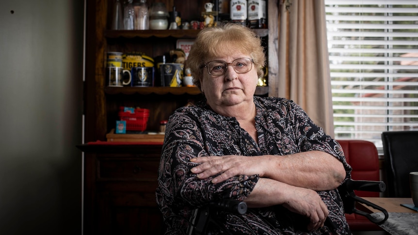 A woman wearing glasses sits at a table with her arms crossed looking at the camera.