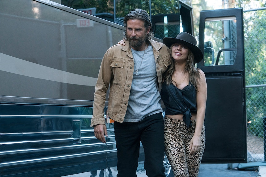 Colour still of Bradley Cooper and Lady Gaga walk together near tour bus in 2018 film A Star is Born.
