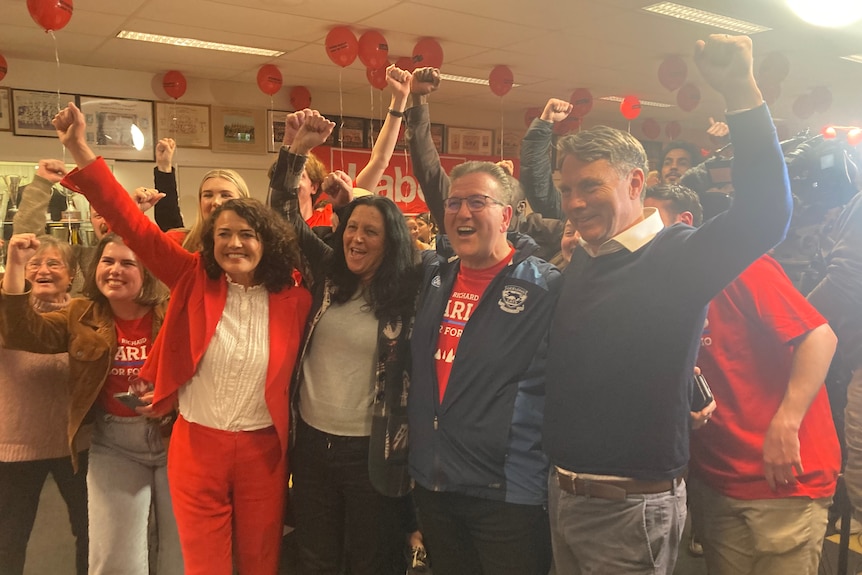 Libby Coker and Richard Marles stand, cheering, with other Labor supporters, in a room decorated in red.